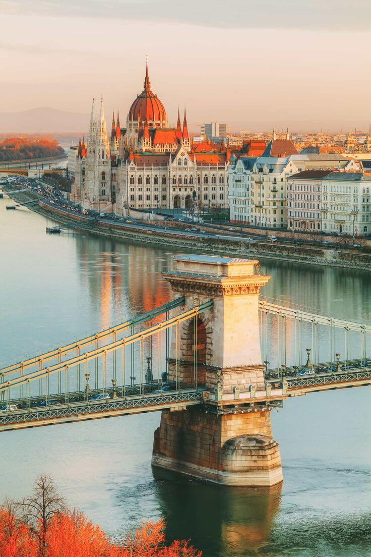 Kota Budapest dengan pemandangan Danube River, jembatan Chain Bridge, dan Parliament Building yang megah di latar belakang, dengan cahaya matahari sore yang memperindah suasana kota